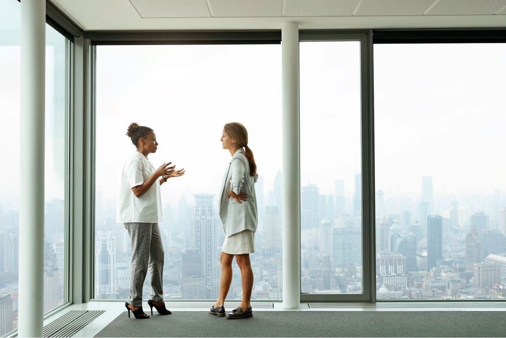 Two women converse in front of a large window showing a city skyline
