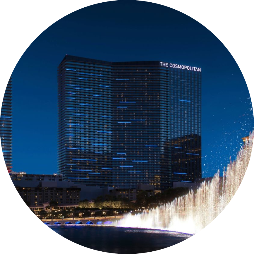 The image features The Cosmopolitan hotel, clearly identified by illuminated signage on the building. The scene is set at night, with the hotel’s modern glass facade reflecting city lights. In the foreground, a dramatic water fountain display is visible, adding dynamic movement and visual interest to the urban landscape.