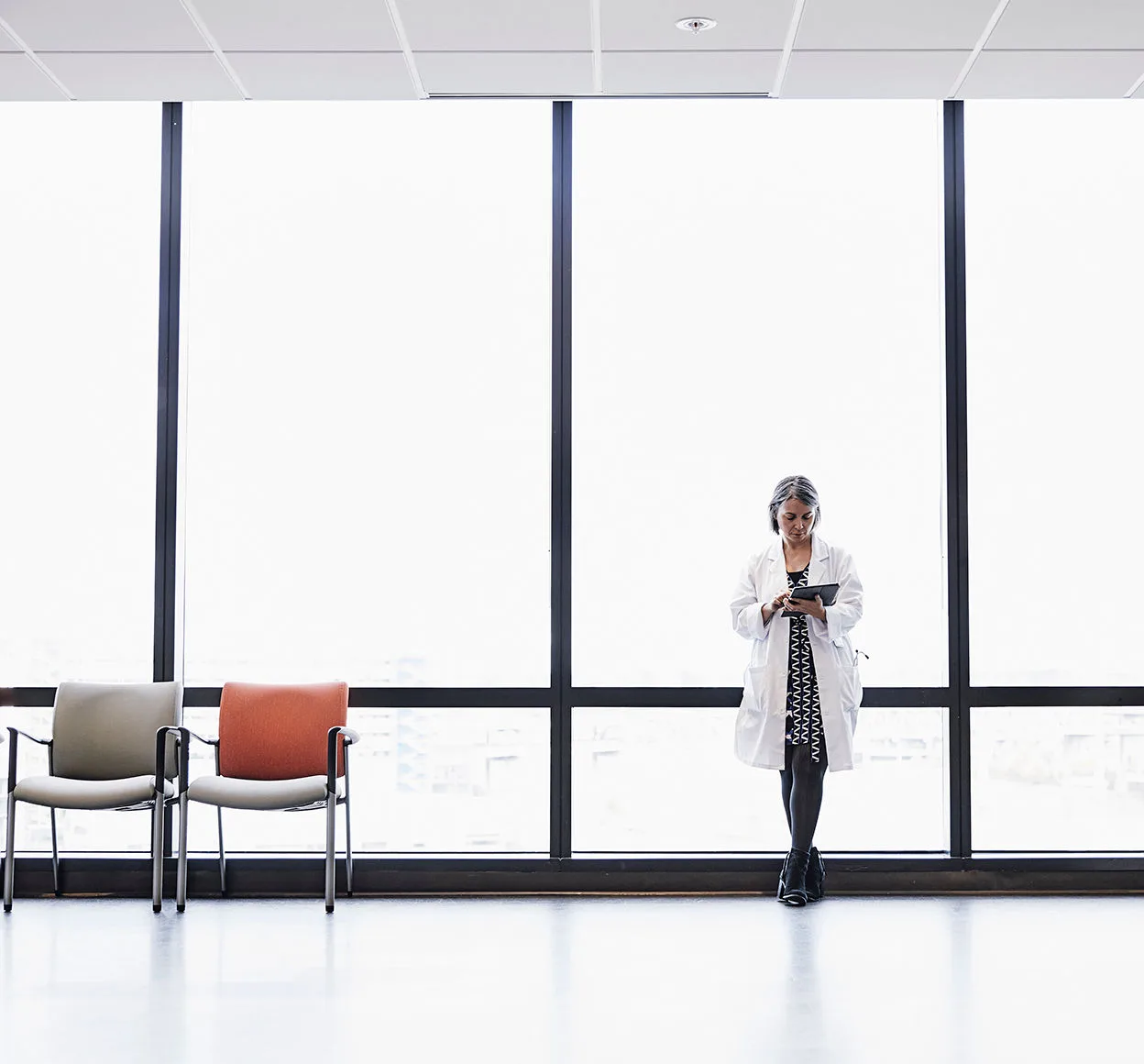 A woman in a lab coat leans again a large office while on a tablet in an empty waiting room.