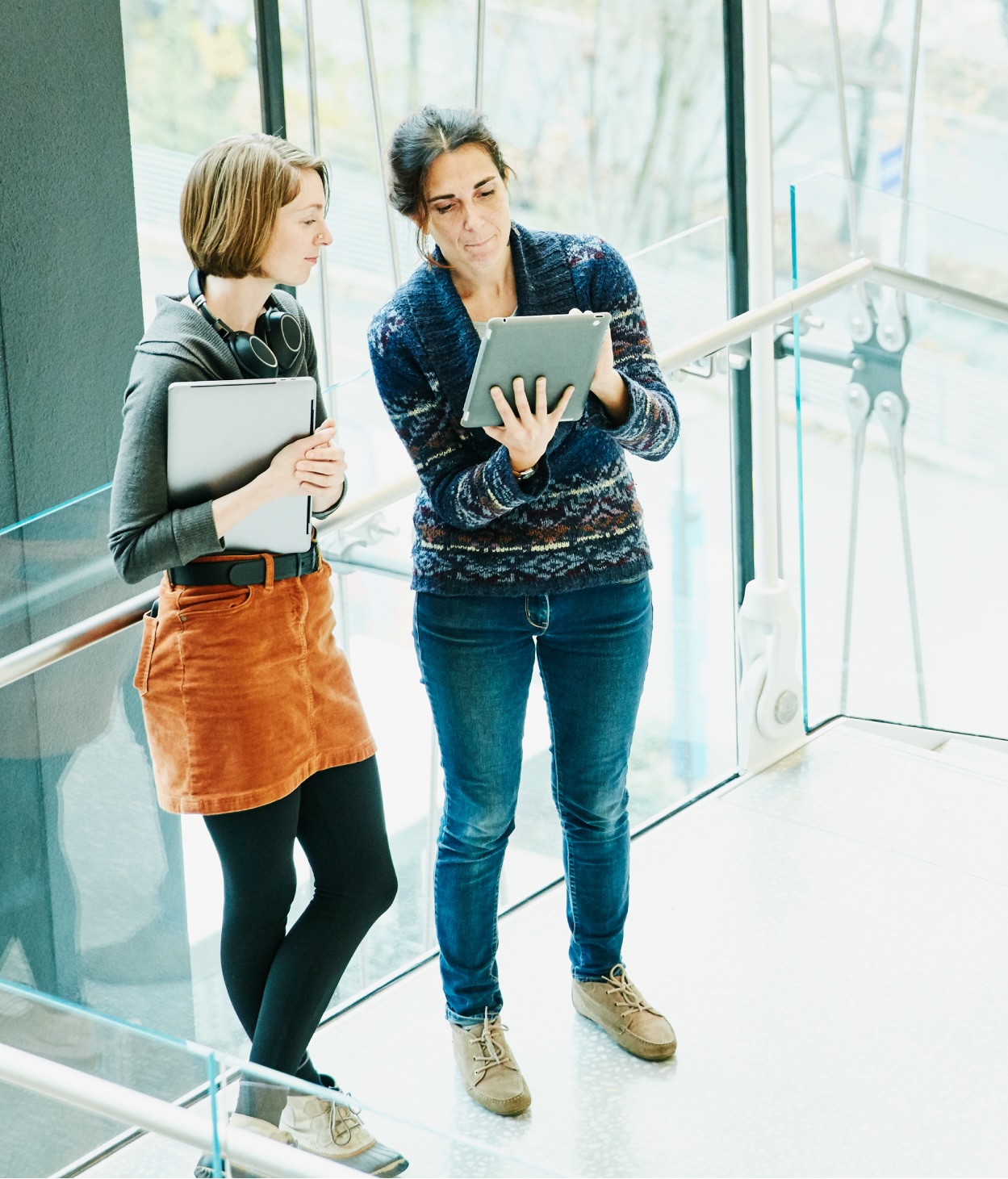 Two women examine a document on a mobile tablet device while on an office walkway.