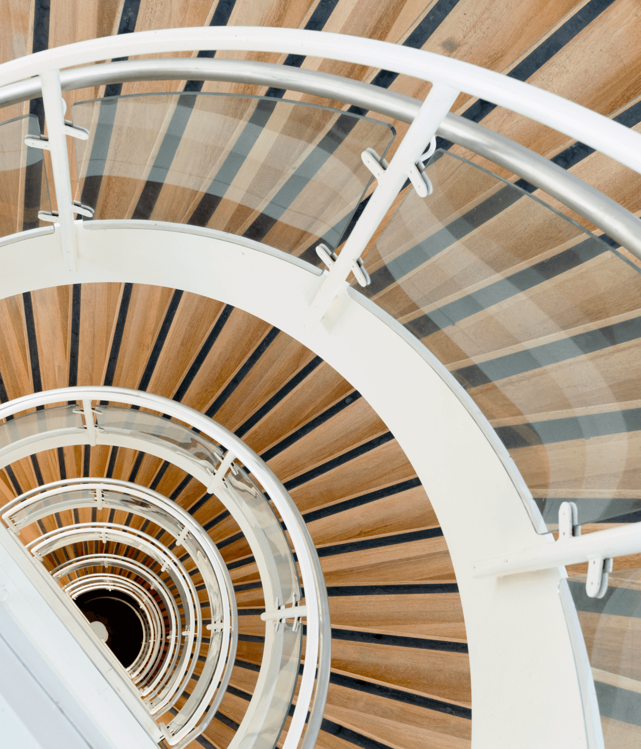 photo of a spiral staircase with glass railing and white metal barristers. The viewer is looking down from the top of the staircase so that the steps form a semi circle.