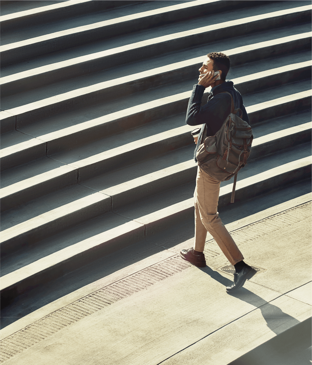 photo of a man in his 20s with a backpack walking up a set of stone stairs outside while talking on a phone.