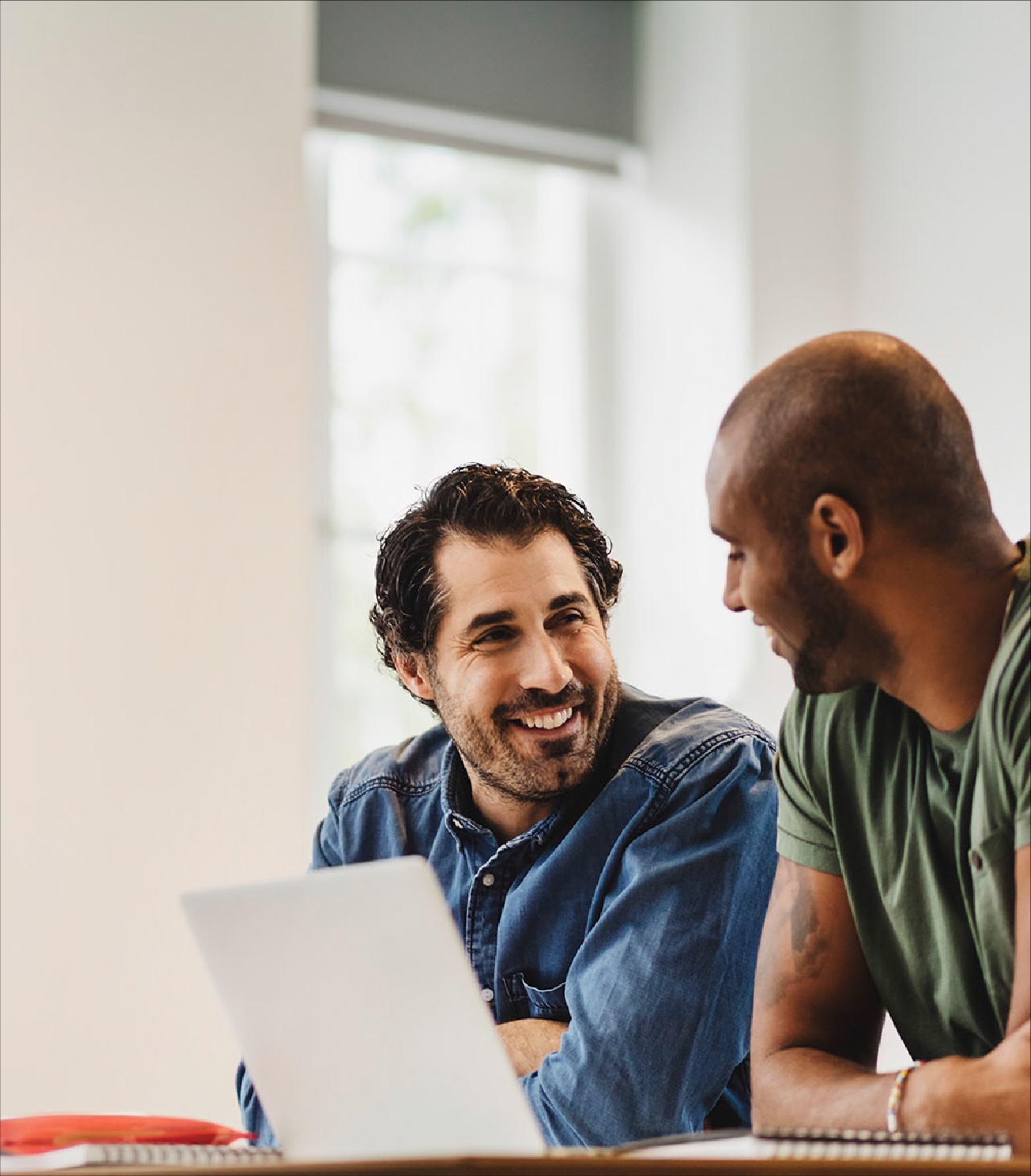 Two coworkers collaborating at a desk, reviewing content on a laptop in a bright office setting.