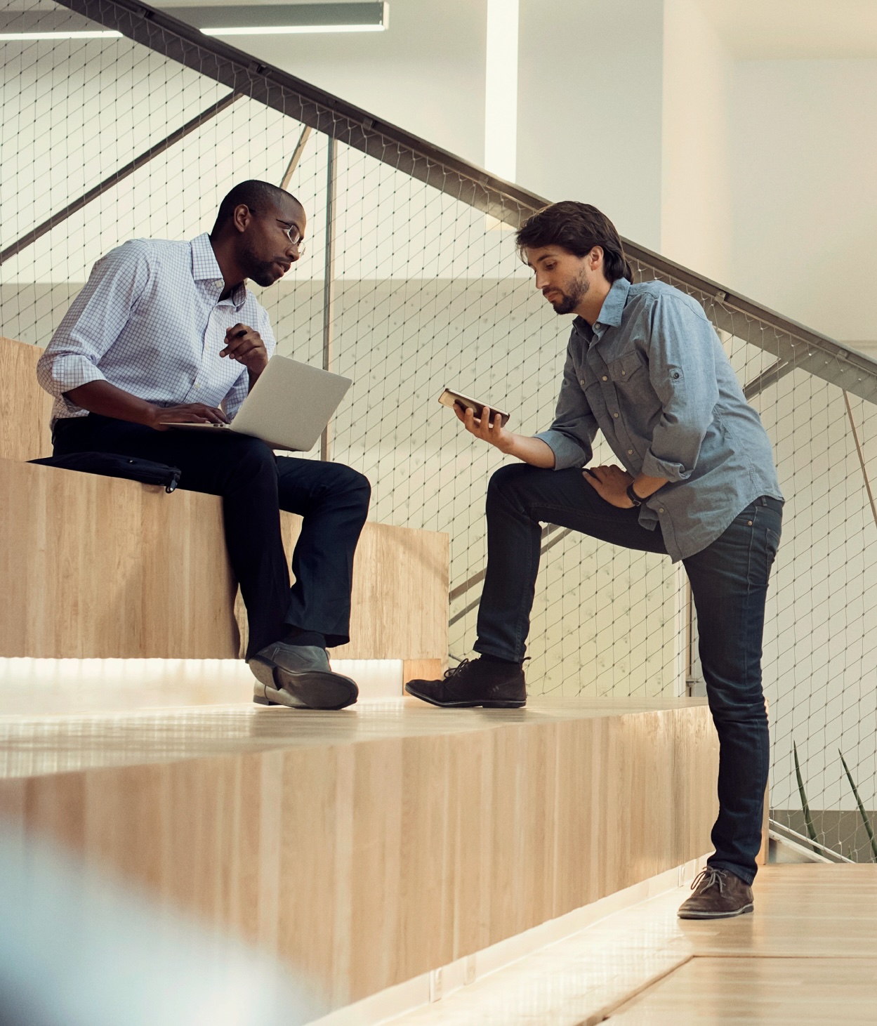 Two businessmen, one with a laptop and the other with a mobile phone, collaborate while sitting on a set of office auditorium steps.