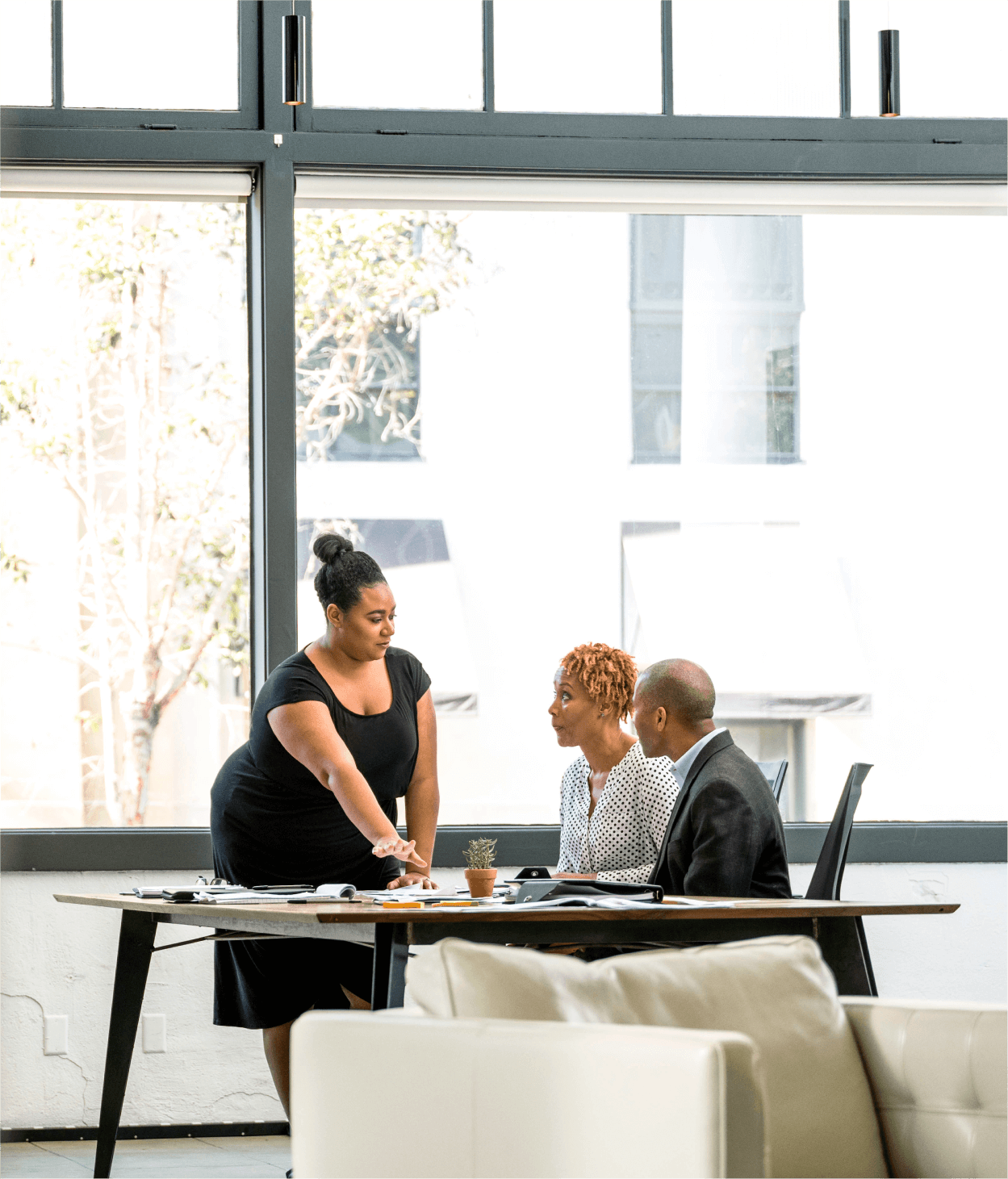 photo of two women in their 20s and a man in his 30s hold a meeting in an office in front of an open window and a white chair.