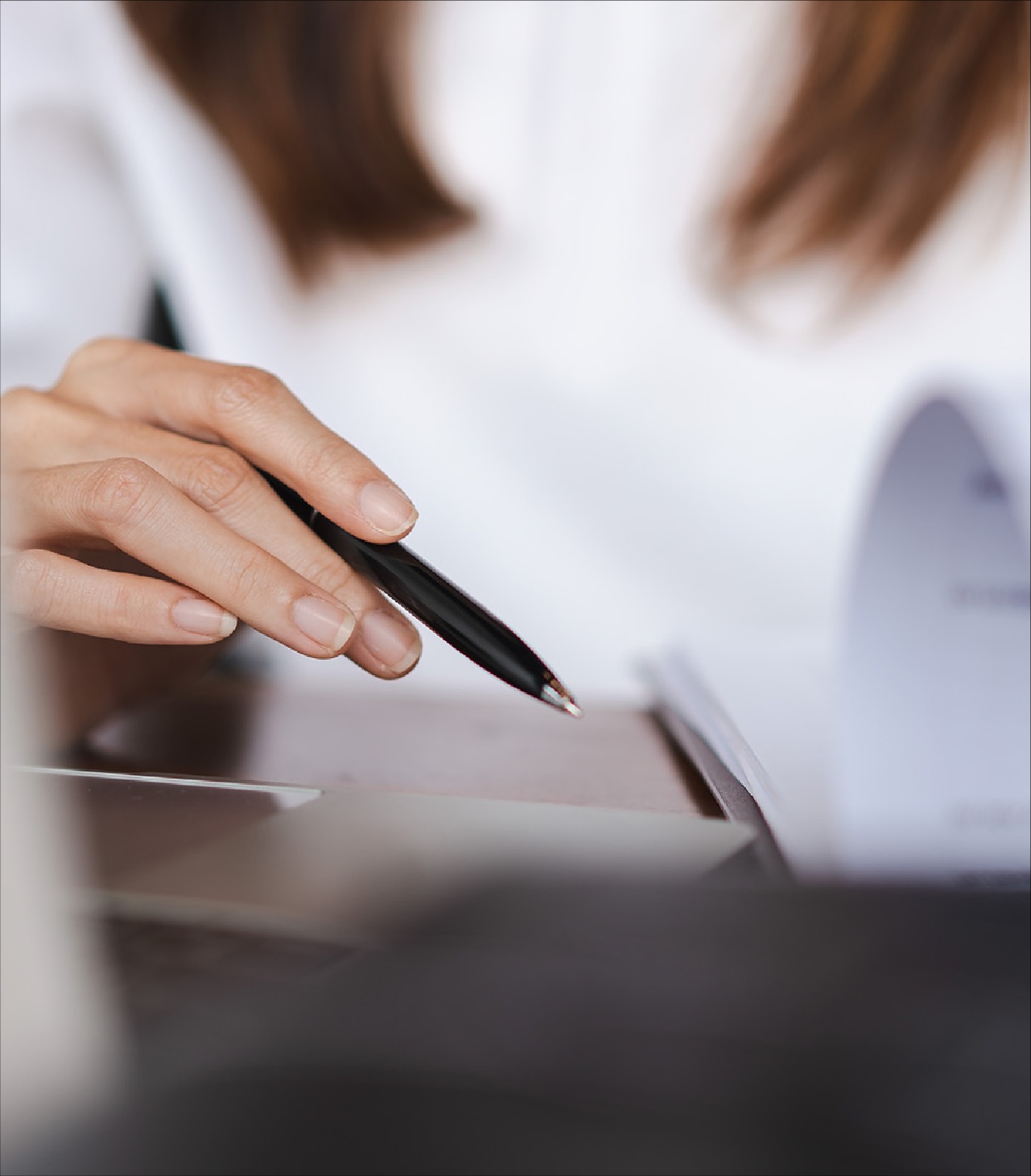 Close-up of a person’s hand holding a black pen and writing on paper at a desk, with an open document blurred in the background.