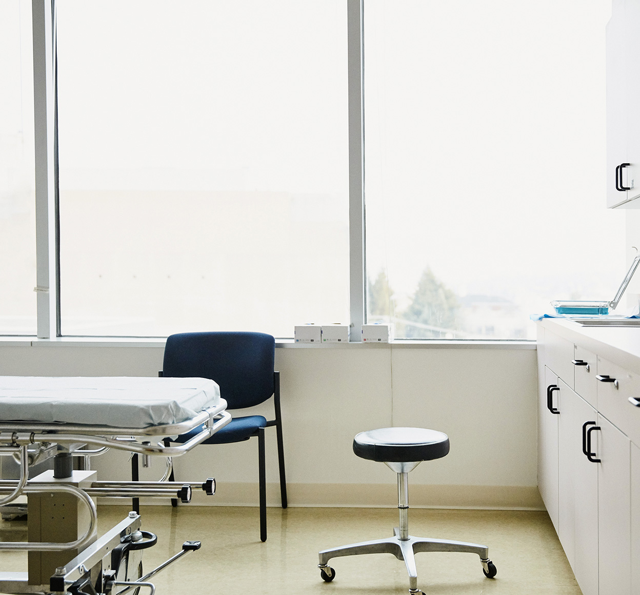 A medical exam room with a stool, patient bed, and large windows