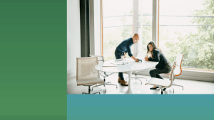 One male and one female employee in their 30s discuss a work project at a table in their office.